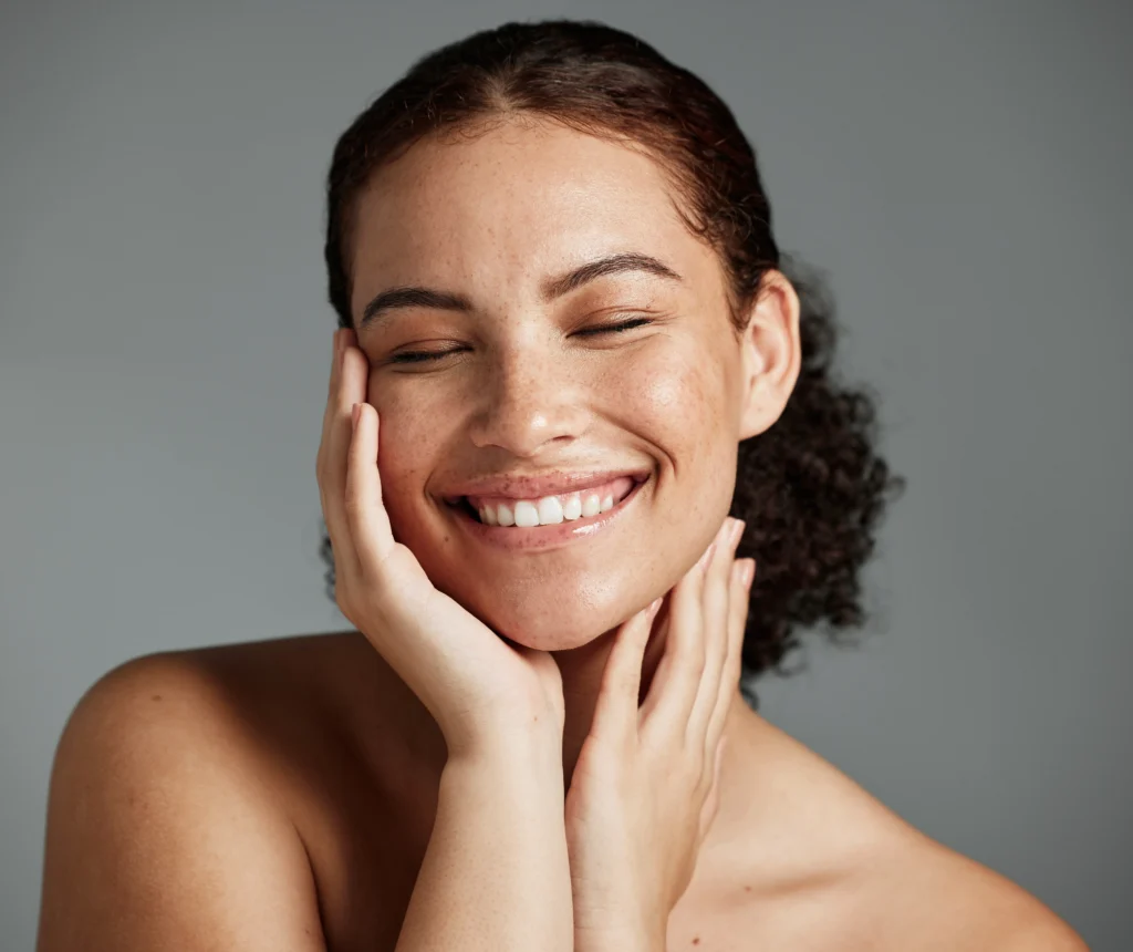 Person smiles with eyes closed while gently touching both sides of the face against a gray background.