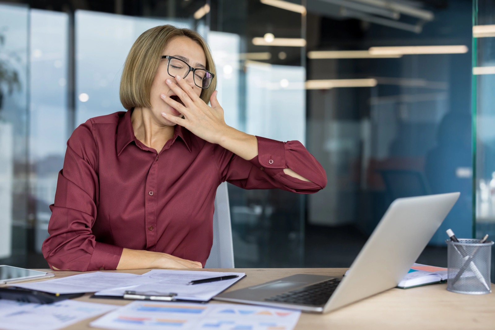 Woman yawning at a desk beside a laptop and paperwork in an office.